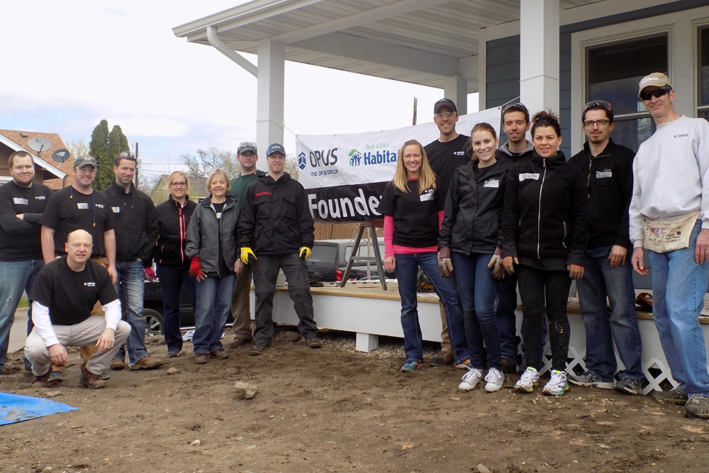 group of men and women in front of banner on house porch that says, "Founder's Day" with The Opus Group and Habitat for Humanity logos